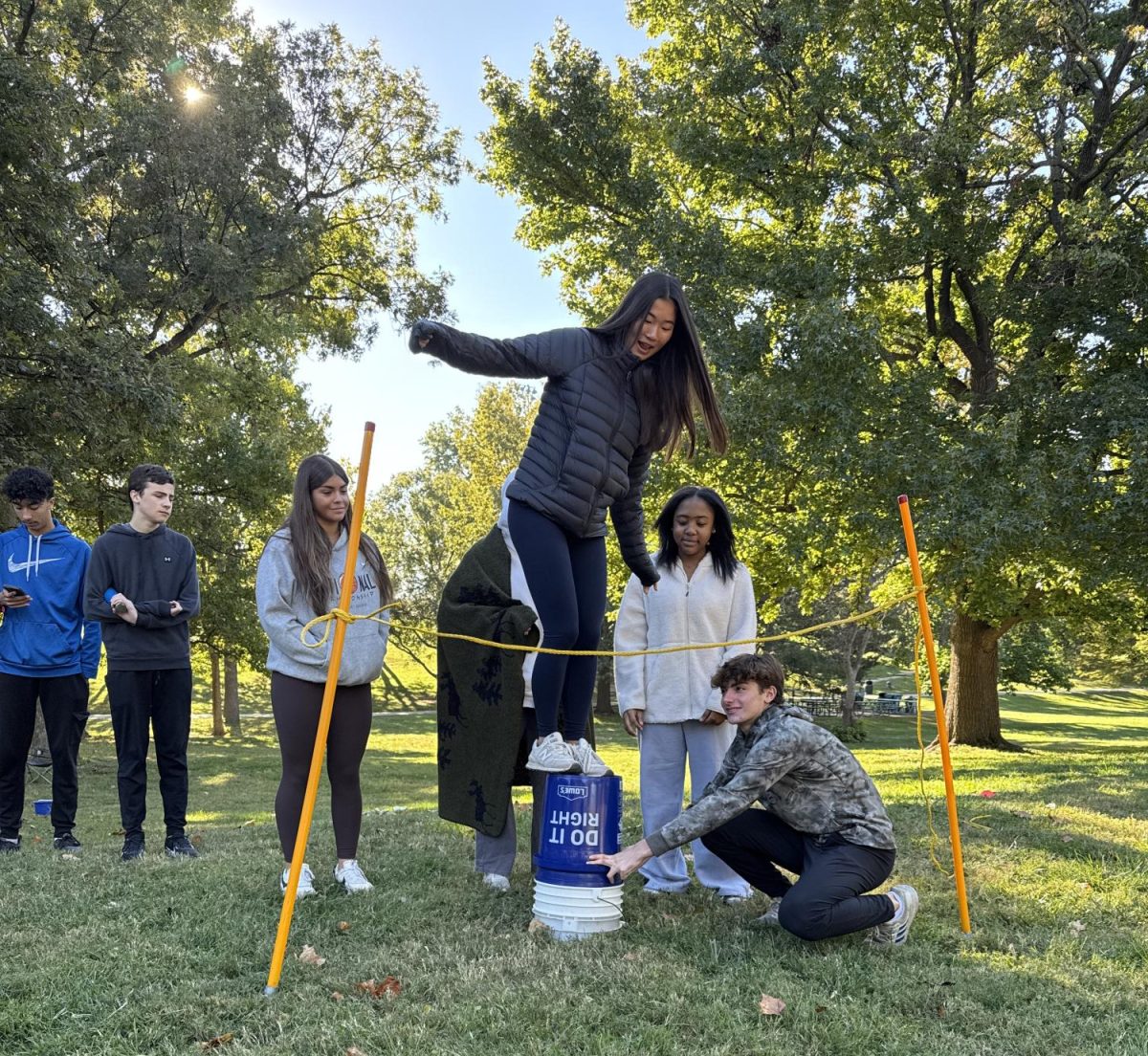 On Wednesday, Oct. 16, the sophomore class gathered at Shaw Park to participate in the sophomore challenge. Throughout the day, students rotated through team-building stations run by seniors. In this station, the rope represented an “electric fence,” and the sophomores had to find a way to get over the fence without touching it. Sophomore Lydia Lee balances on buckets to get over the obstacle.